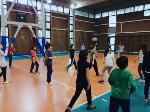 La palestra ha il pavimento in linoleum color arancione e su di esso ci sono le linee bianche che delimitano il campo per la pallavolo. Tutto esterno al campo è di color verde smeraldo. Ci sono dei bambini che stanno giocando.