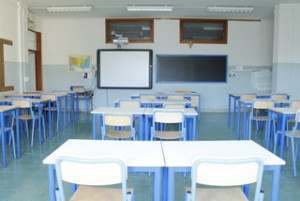 un'aula didattica con banchi e sedie bianchi e blu, lim e pc, lavagna classica nera a gesso, bacheca a parete e armadio per la biblioteca di classe.