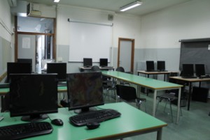 Aula d'informatica con pc neri e schermi piatti, tavoli verdi, aria condizionata e videoproiettore.