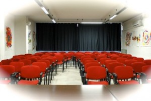 Aula magna: sedie rosse della platea e lato finestrato con tende oscuranti, aria condizionata e quadri realizzati dai ragazzi alle pareti.